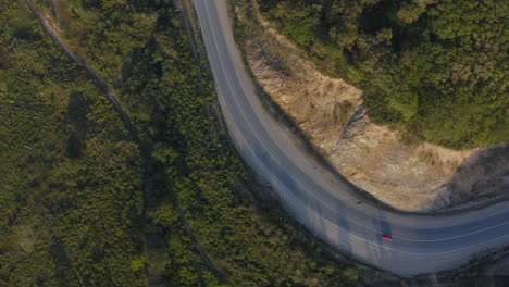 vista superior de un coche de color rojo que pasa por una larga carretera curva en las montañas verdes con un acantilado en el lado izquierdo, durante la puesta de sol