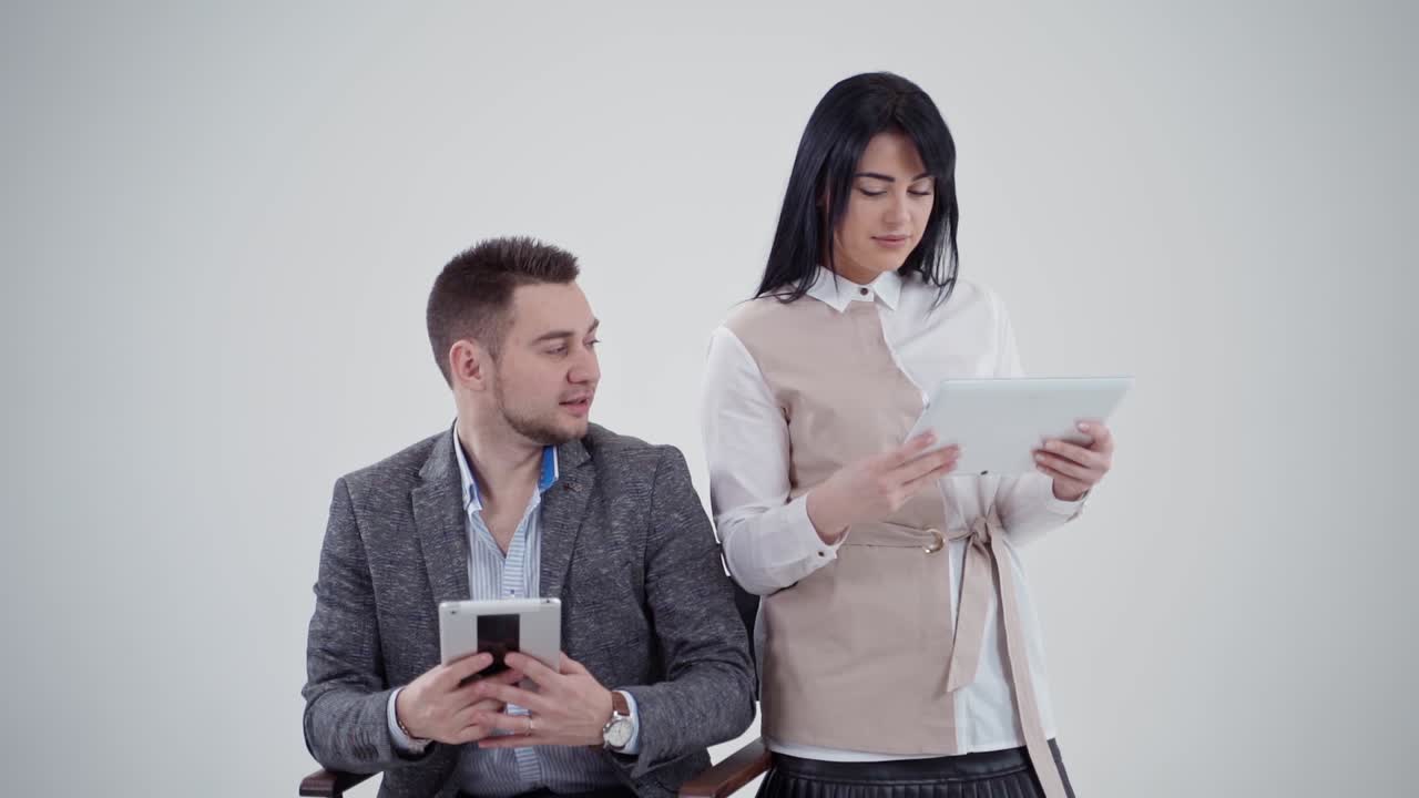 Man and woman are holding gadgets. Handsome man talking to a pretty woman while looking into the tablet isolated in studio. Beautiful lady smiling. Slow motion.