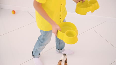 a boy carries two dog bowls, and one of his dogs follows him as he leaves the room