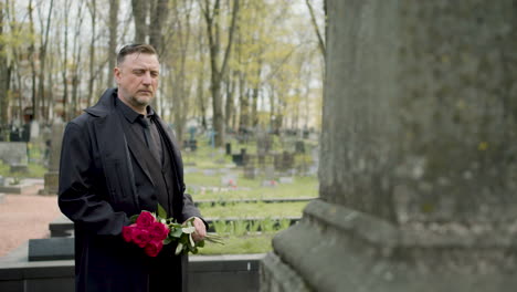 man in black raincoat and suit holding red roses standing in front of a tombstone in a graveyard