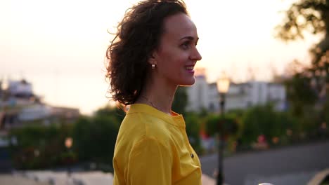 smiling brunette woman in a long yellow dress standing by her city bicycle holding its handlebar with flowers in its basket