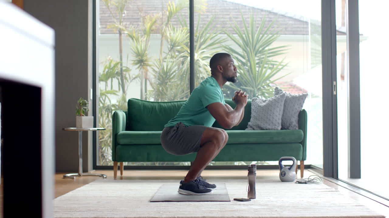 Uomo che si esercita a casa, facendo squat sul tappetino da yoga con un kettlebell nelle vicinanze.