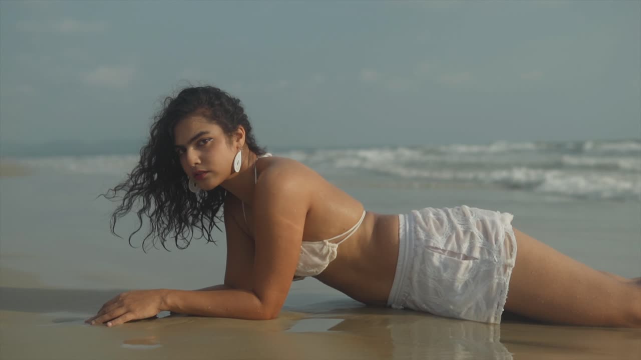 A captivating clip features a beautiful woman in her early 20s, wearing a white bikini top and short white shorts, lying on the wet sand