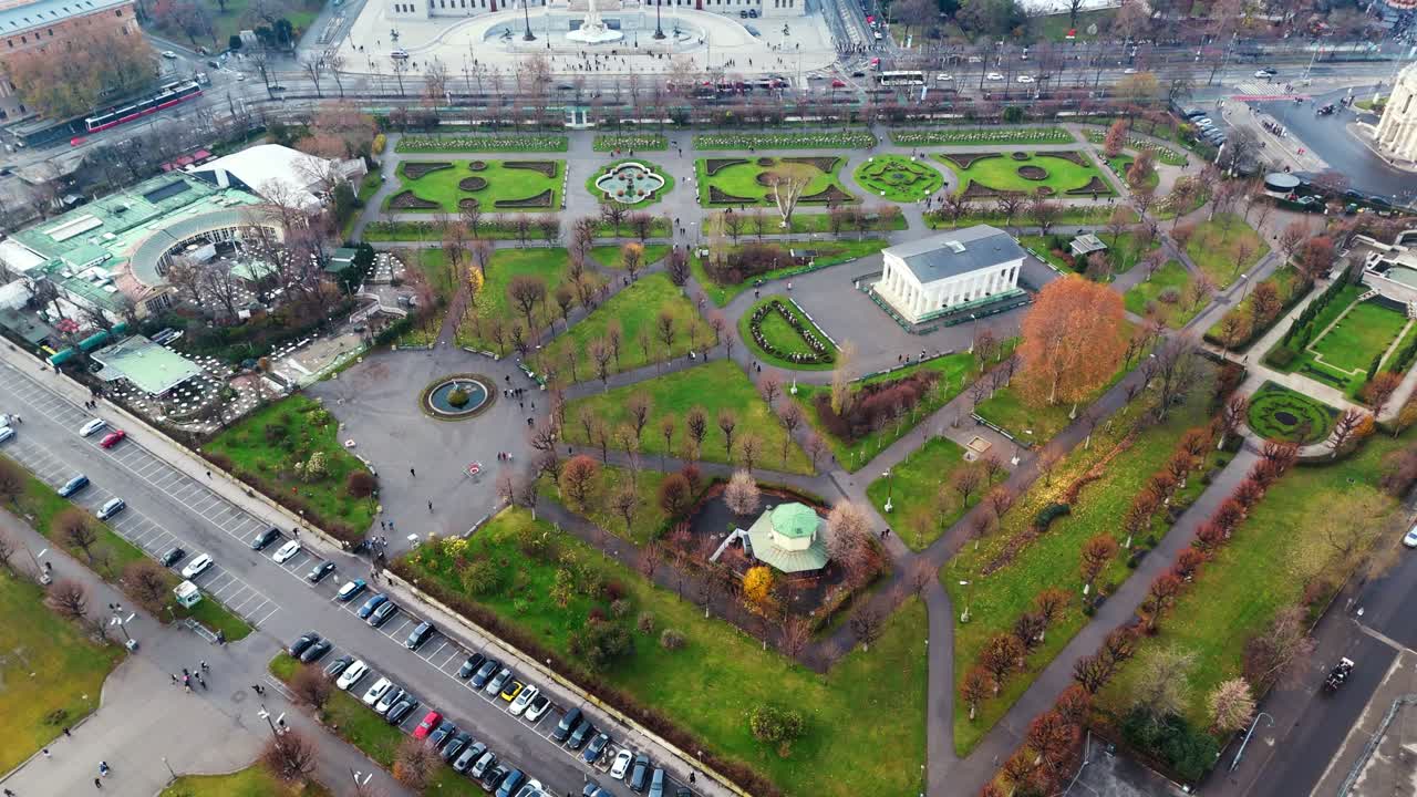 Panoramatický záběr z dronu na zahrady Hofburgu ve Vídni z ptačí perspektivy, ukazující zimní trávu, upravené zahradní cesty, okolní palácové budovy, jasné zimní světlo a turisty