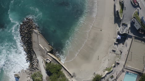 4K-Kinoaufnahme-Von-Oben-Vom-Strand-Lovers-Point-Mit-Wellen,-Die-über-Die-Felsen-Des-Wellenbrechers-Schlagen