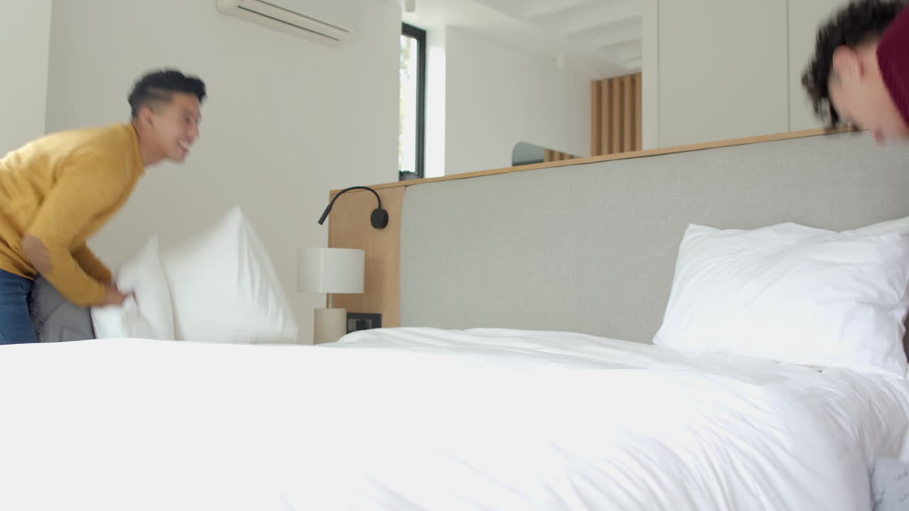 Having fun, multiracial gay couple enjoying playful pillow fight in modern bedroom, at home