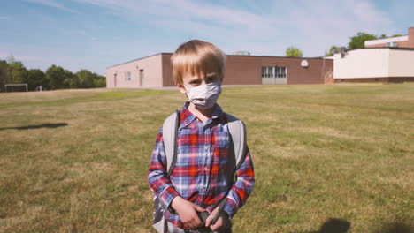 little boy in a face mask turns and walks toward school