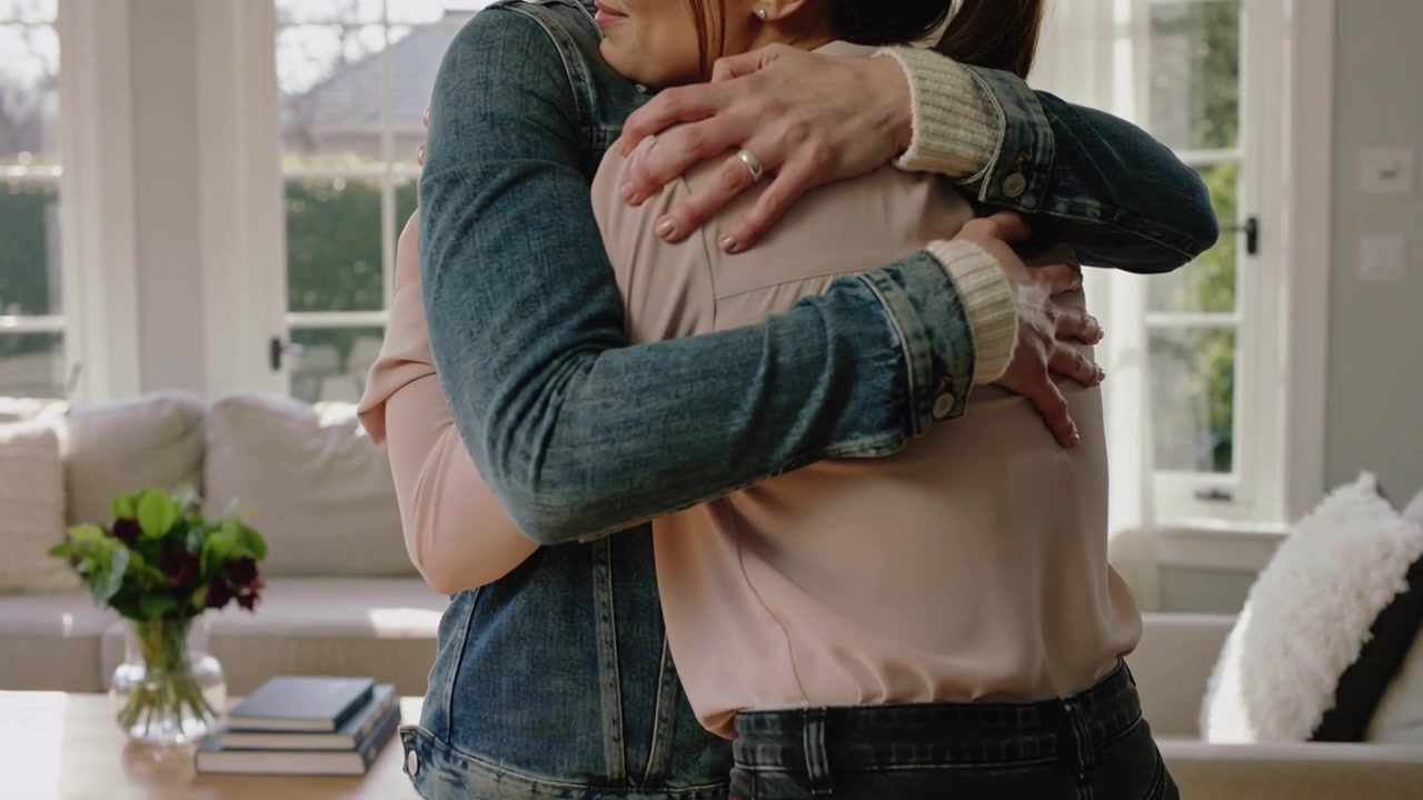Stepping forward two women embracing in greeting at home, denim jacket, pink blouse, ring visible