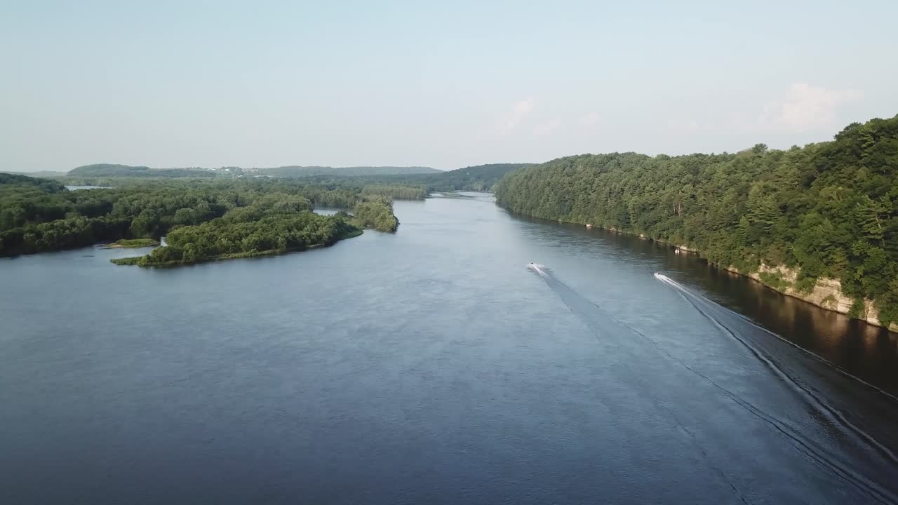 Twin speedboats leave crisp wakes as they dash upriver between densely wooded hills, with green forest edges framing a smooth waterway on a clear summer day.
