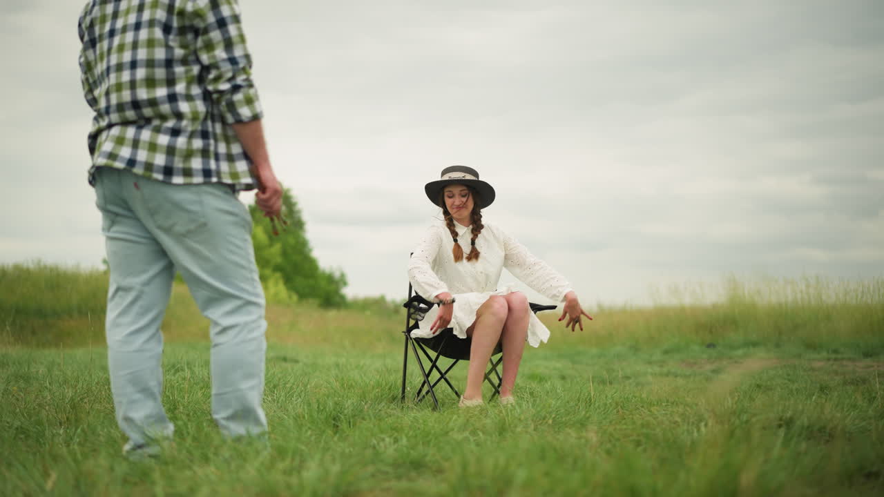 하 드레스와 검은 모자를 입은 행복한 여자가 잔디에 자리를 잡고 의자에 앉으려고 준비하고 있습니다. 색 셔츠를 입은 남자가 을 들고 예술적인 의도로 그녀를 관찰합니다.