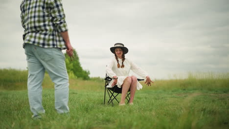a happy woman wearing a white dress and black hat confidently poses in a grassy field, preparing to sit on a chair. a man in a checked shirt, holding a brush, observes her with artistic intent