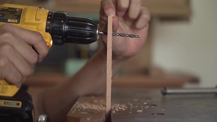 Closeup of craftsman drilling wooden strip with cordless drill, creating precise hole while supporting timber during woodworking process