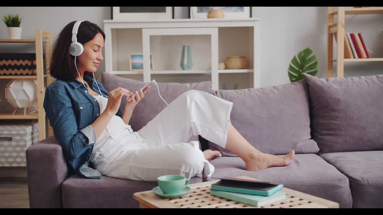 여성이 소파에서 휴식을 취하고 음악을 듣고 전화를 사용하고 있습니다.