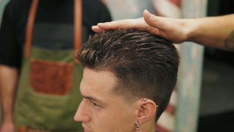 vista de cerca de un joven apuesto hombre caucásico con perforación en la oreja recibiendo su cabello vestido y estilizado por un barbero barbudo