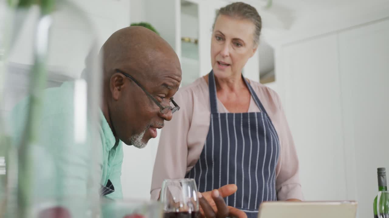 Couple senior mixte souriant portant des tabliers bleus et utilisant une tablette dans la cuisine.