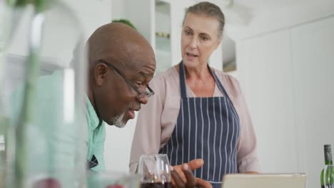 una pareja diversa y sonriente con delantales azules y usando una tableta en la cocina