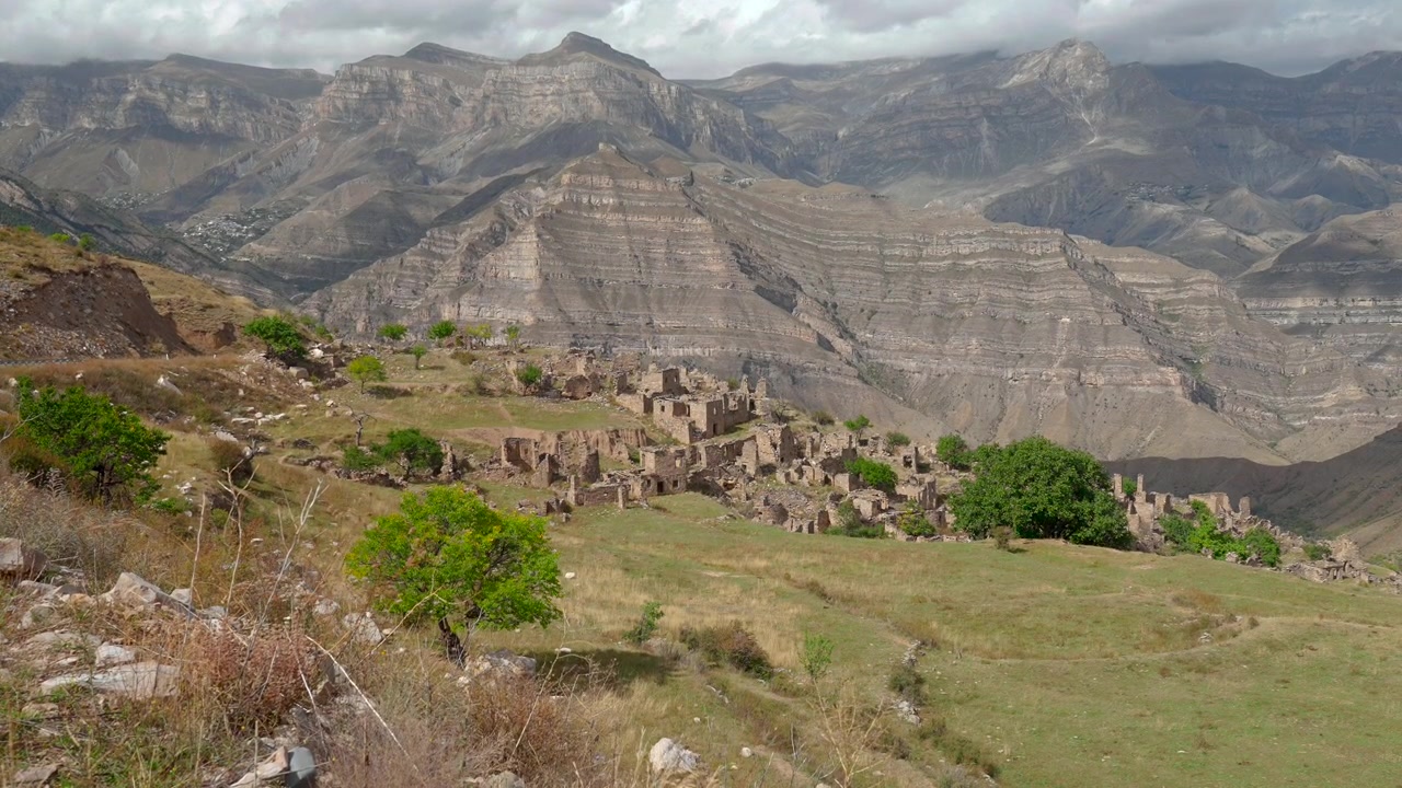 Ampia vista dall'alto mostra un insediamento in pietra in rovina su un pendio erboso e secco, incorniciato da scogliere rocciose, alberi sparsi e luce tenue e coperta attraverso un'ampia valle di montagna, creando un tranquillo paesaggio storico.