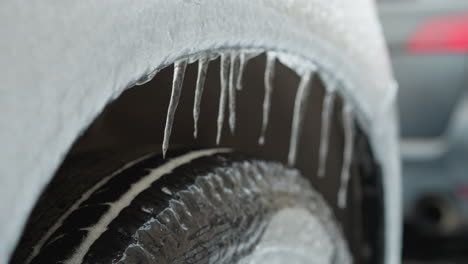 close-up of car tire covered with snow and hanging icicles during winter, featuring a blurred background with the rear view of another car