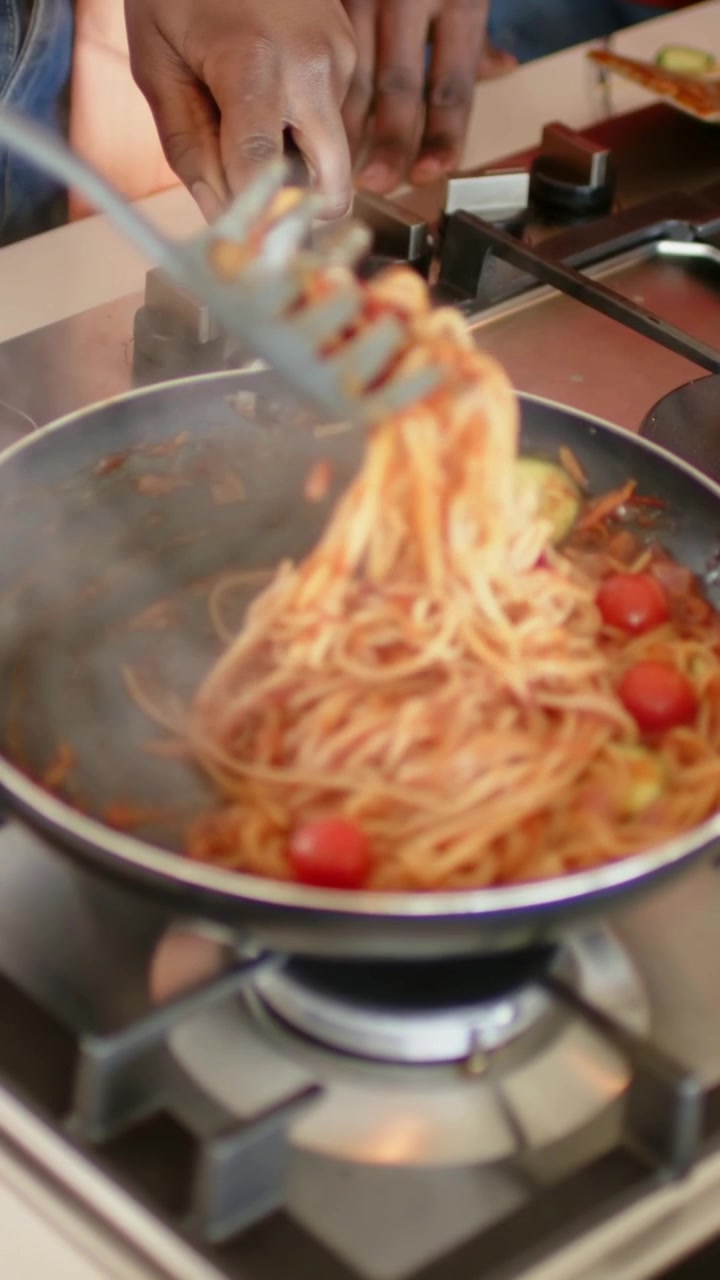Video verticale: Un cuoco che prende un mestolo, mescolando per condire i noodles su un fornello a gas, con i pomodori.