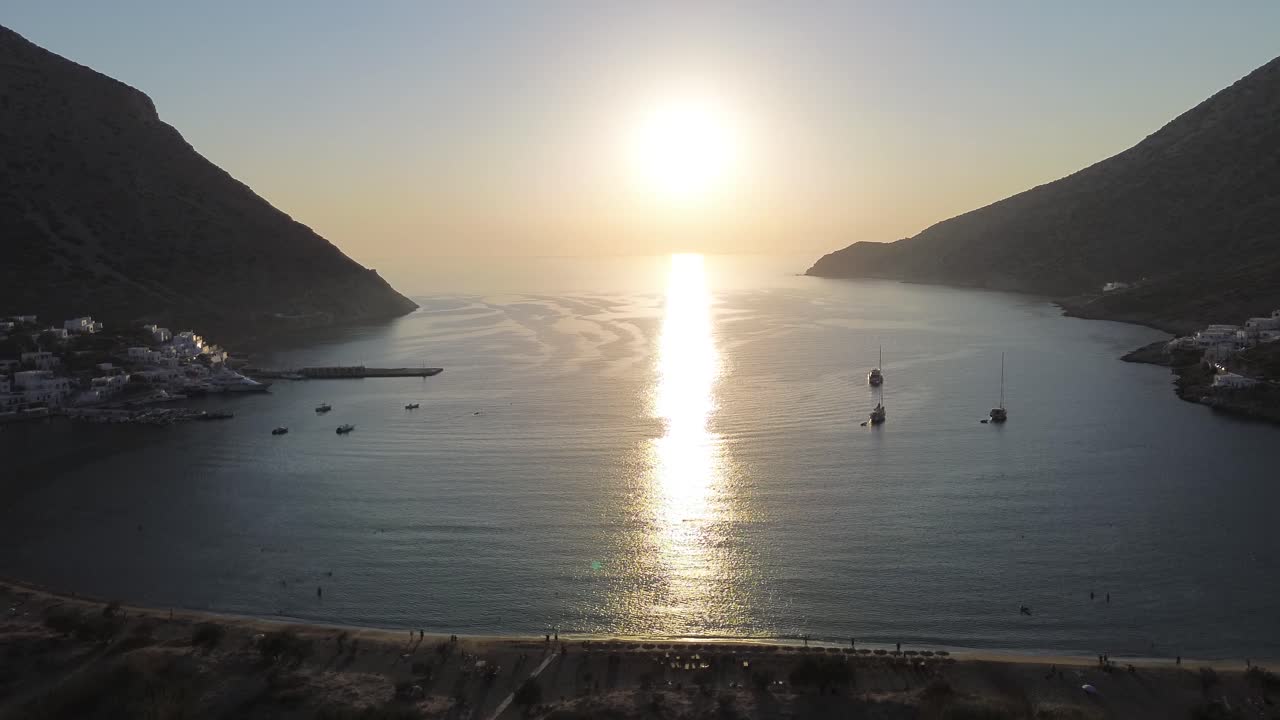 Tomada cinematográfica de drone em ângulo elevado do pôr do sol entre as montanhas da baía de Kamares em Sifnos, Grécia, com reflexos dourados no mar e a vila costeira em vista