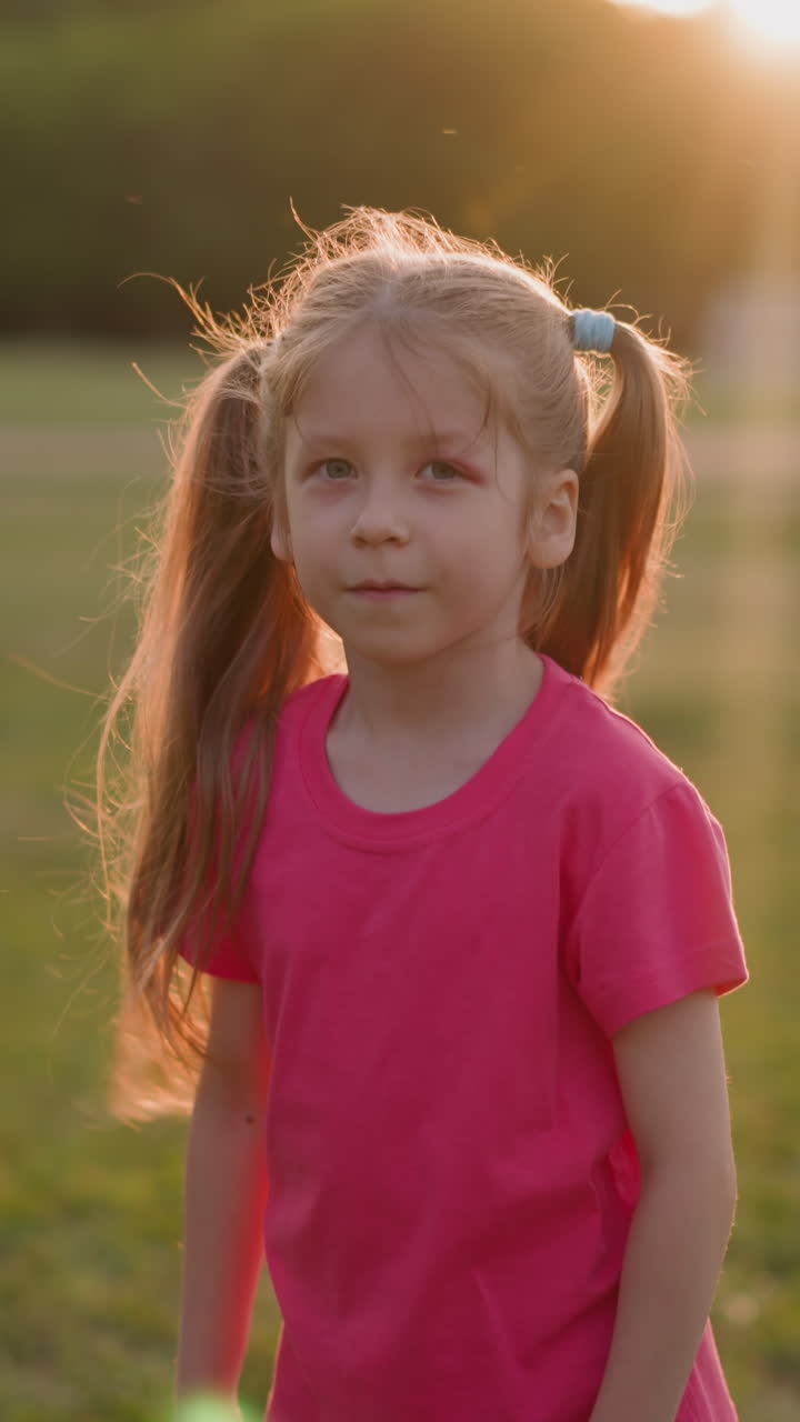Adorable little girl with wounded eye shakes hair and looks in camera on playground at sunset slow motion. Public place for rest and activity