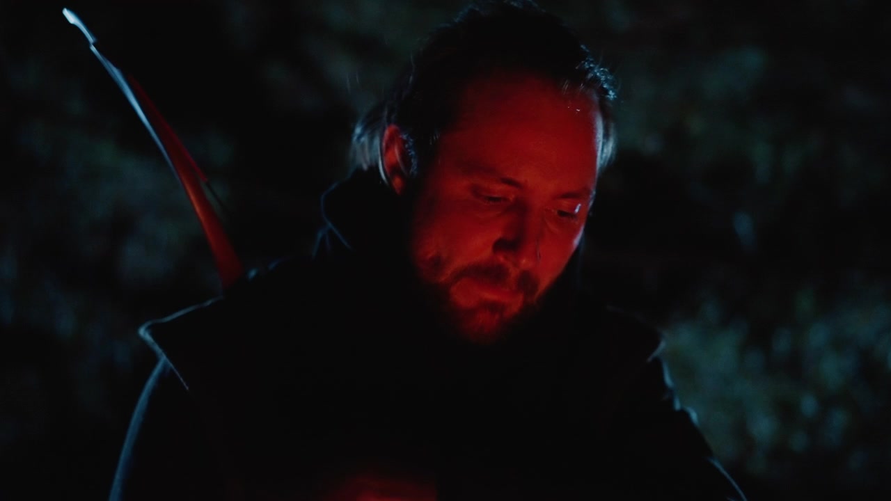 Un uomo incappucciato con arco lungo abbassa la testa e guarda in basso sotto un'illuminazione notturna rossa e blu, mostrando una concentrazione stanca, una tensione silenziosa, dettagli scuri del costume e un'atmosfera cinematografica del personaggio. Arciere.
