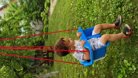 Adorable toddler swaying in the blue swing. Happy time of a beautiful baby boy outdoors in summer. Vertical screen.