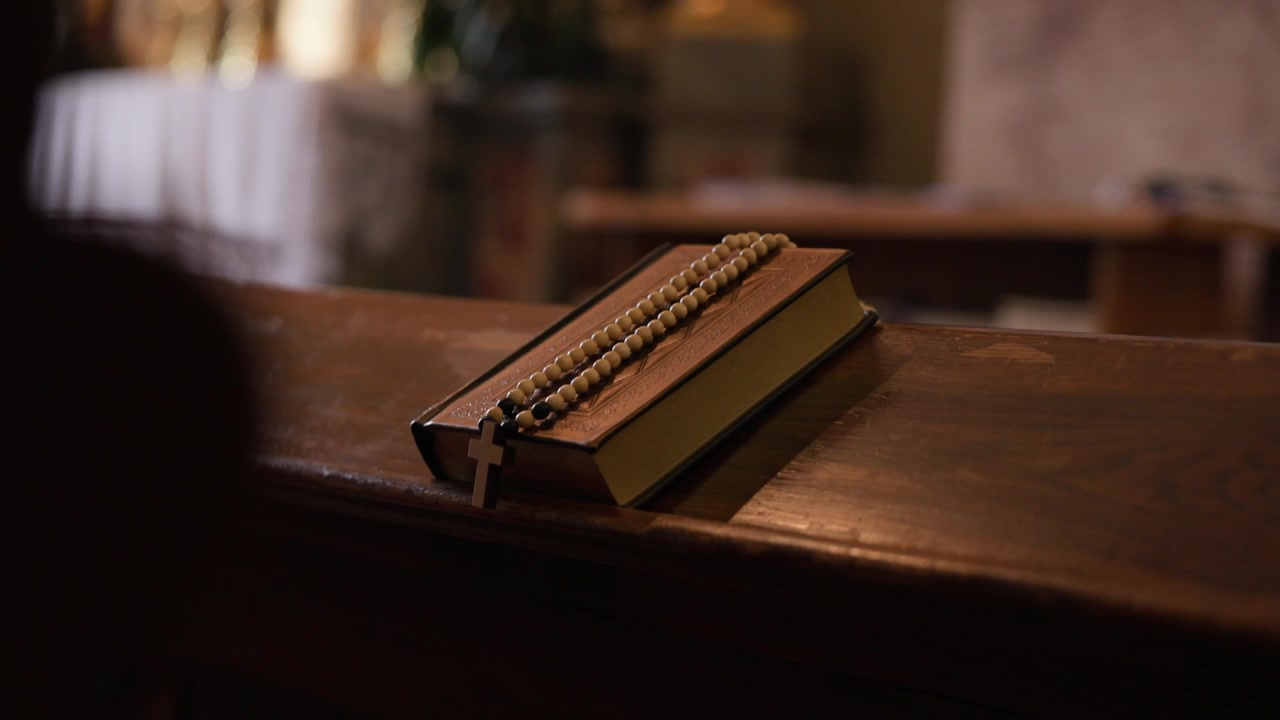 Bibbia cattolica e rosario con croce poggiati su una panca di legno di chiesa che simboleggiano la fede cristiana.