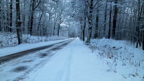 Low-angle-drone-flying-along-the-snow-covered-and-ice-covered-countryside-forest-road,-huge-snowdrifts-on-sides