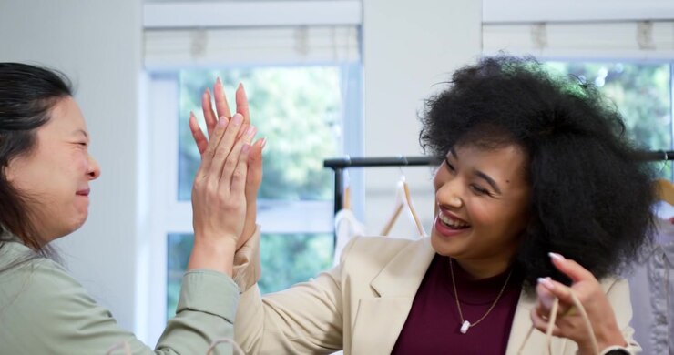 Diverse female friends finding items in brown paper bags at boutique rack, celebrating, high-fiving