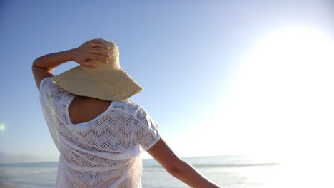 Una-Joven-Birracial-Disfruta-De-La-Playa-Con-Espacio-Para-Copiar,-Protegiéndose-Los-Ojos-Con-Un-Sombrero-De-Ala-Ancha