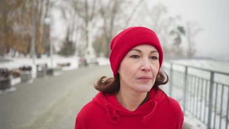 mujer corriendo, mirando cansada a lo largo del camino nevado del parque en sudadera roja con capucha y leggings negras, rodeada por la atmósfera de invierno brumosa con bancos, arbustos, barandillas de hierro y postes de lámparas detrás