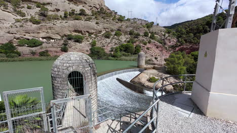 A-Reservoir-Dam-With-Water-Gushing-Down-Near-A-Rocky-Mountain-With-Green-Trees-And-Shrubs