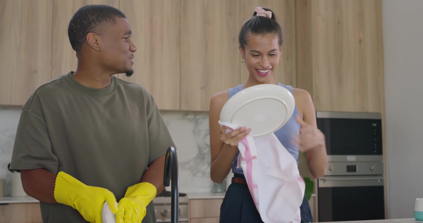 Couple at sink washing dishes after meal, man in gloves rinsing, woman drying with striped towel