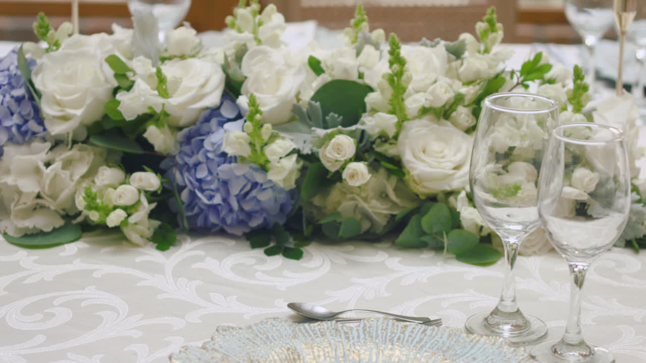 A detailed clip of an elegantly decorated wedding table. It features stemware, a white tablecloth, plates, cutlery, cloth napkins, a delicate floral arrangement, and a personalized invitation.