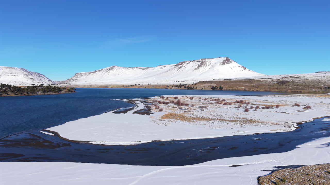 Drone backward shot over Caviahue lake with snowy mountains, frozen ground, and a bright blue sky in Patagonia, Argentina