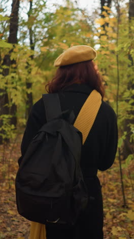 vista trasera de la dama con abrigo negro, boina amarilla y mochila caminando a través de un tranquilo parque de otoño con árboles altos y follaje seco, ajustando su silenciador de cuello mientras disfruta de un retiro pacífico en la naturaleza