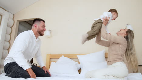 loving parents play cheerfully on the bed with their baby boy. dad tosses a kid to mom's hands. low angle view.