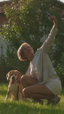 woman taking selfie with her dog focused on something else outdoors in lush green garden under sunny weather, with building and trees in background, dog is sitting, panting, while owner smiles