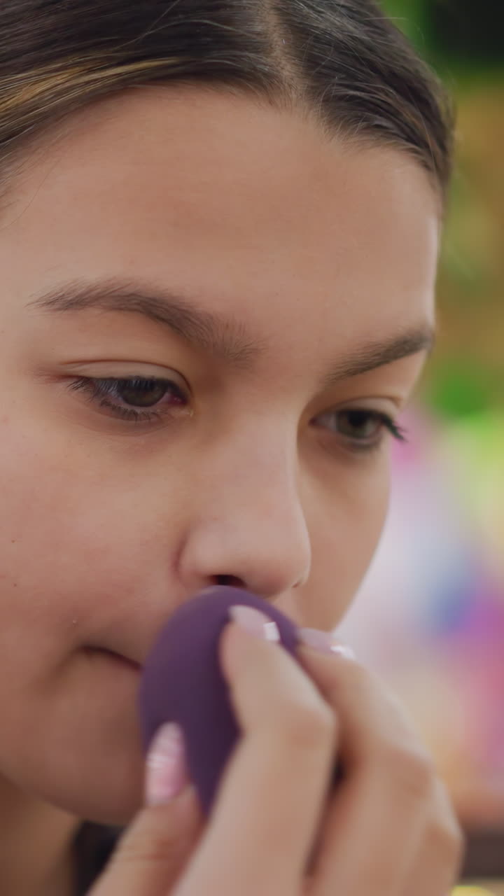 Primer plano de una adolescente aplicándose maquillaje con una esponja; modelo pensativa examinando su piel en el espejo mientras difumina la base y el corrector con una esponja morada, luz natural suave, tonos neutros, uñas arregladas, calma
