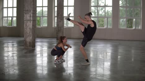 beautiful young woman with dreadlocks doing squats exercise with man partner lifting legs under her head synchronously. fitness loft studio with big windows on background