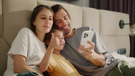 A-brunette-man-in-a-gray-T-shirt-takes-a-selfie-with-his-wife-and-little-brunette-daughter-in-a-yellow-dress-using-a-white-phone-on-a-light-brown-bed-in-a-modern-apartment