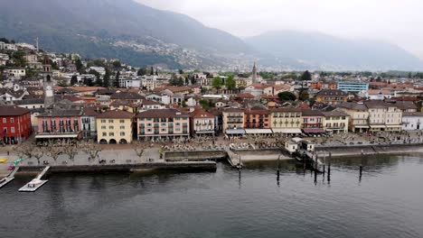 aerial flyover along the promenade of the city of ascona in ticino, switzerland at the shores of laggo maggiore with a side view of colorful houses, rooftops, and boats on a cloudy spring day