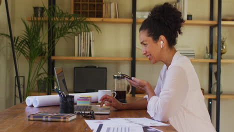 mujer de raza mixta usando una computadora portátil y hablando en un teléfono inteligente mientras trabaja desde casa