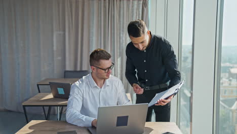 male employees discussing work issues in light office. man in dark clothes shows papers to his colleague sitting in front of laptop.