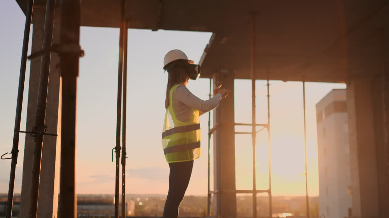 A woman engineer at a construction site in virtual reality glasses moves her hands simulating the work of the interface of the future at sunset