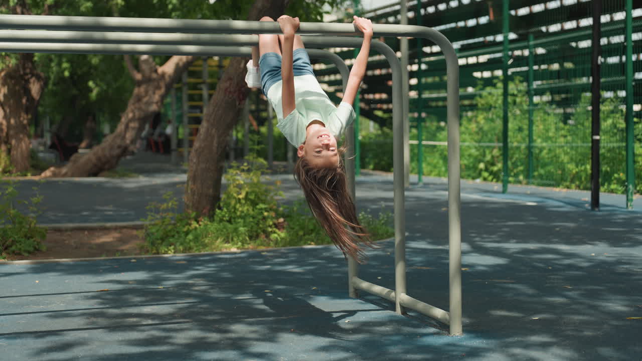 Daughter hanging on playground metal bars, denim shorts and braided hair swinging, sun dappled rubber surface, joyful expression, energetic movement, summer park scene, confident play, childhood