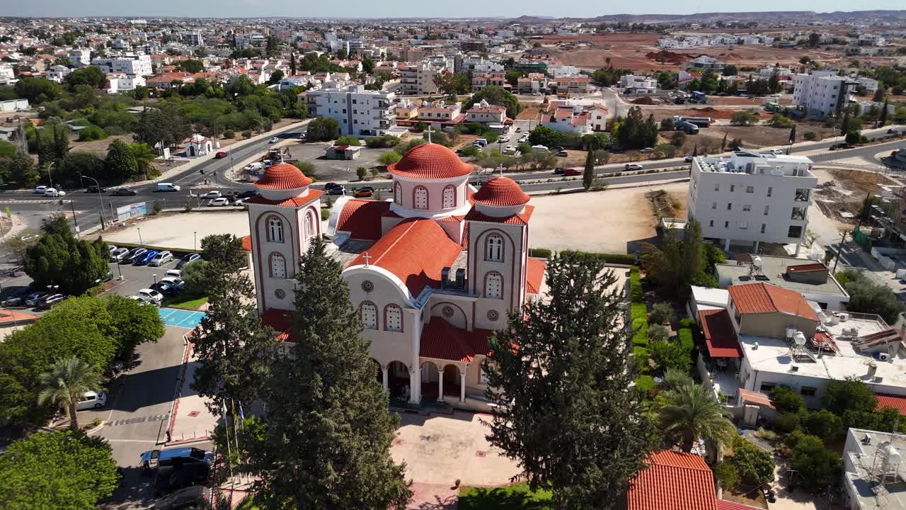 This stunning aerial view captures the church with its red-tiled roof surrounded by green trees and a vibrant neighborhood in Cyprus. The bright sky and landscape add to the charm