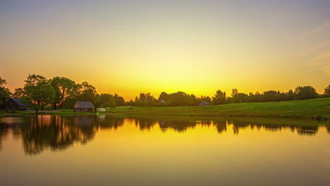 Timelapse-shot-of-sun-rising-over-small-cottages-along-lakeside-surrounded-by-lush-green-grasslands-during-morning-time