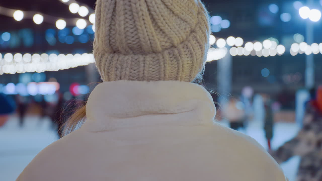 Close-up of woman in warm winter beanie standing still while people skate and move around under glowing decorative lights, with illuminated Christmas tree and blurred city background